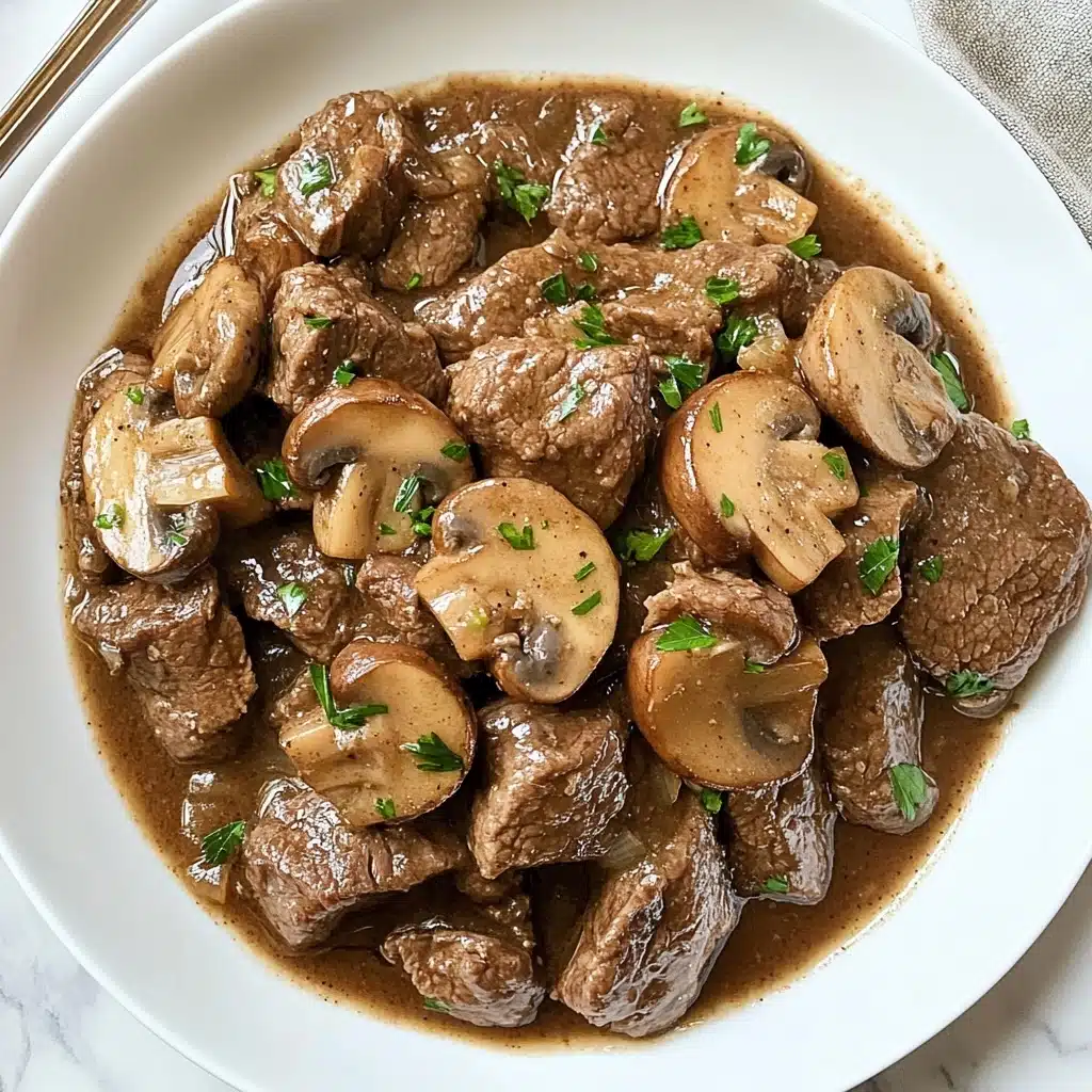 Instant Pot Garlic Butter Steak Bites, quick beef recipes, how to make steak in Instant Pot, tender steak bite ideas, easy Instant Pot dinner A white plate holds a dish with many pieces of cooked brown beef tenderly mixed with light brown mushroom slices. The beef and mushrooms sit in a glossy brown sauce that fills the plate base. Small green parsley bits are scattered on top, adding bright spots of color. The plate is placed on a white marbled texture surface with part of a silver fork visible on the left side. The overall look is warm and hearty, with a natural, simple presentation.