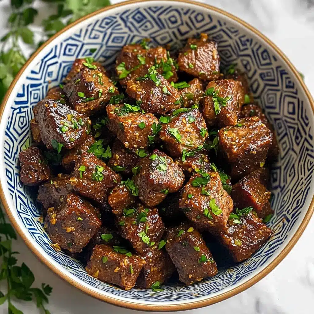 Garlic Butter Steak Bites, Quick Steak Bites, Easy Steak Dinner, Juicy Steak Bites, Tender Steak Appetizer A bowl filled with many small, dark brown cooked meat cubes that have a slightly crispy texture on the outside, sprinkled with chopped green herbs. The bowl is white with blue geometric patterns inside and round brown dots near the edges. The background is a white marbled surface with some green leaves on the side. The