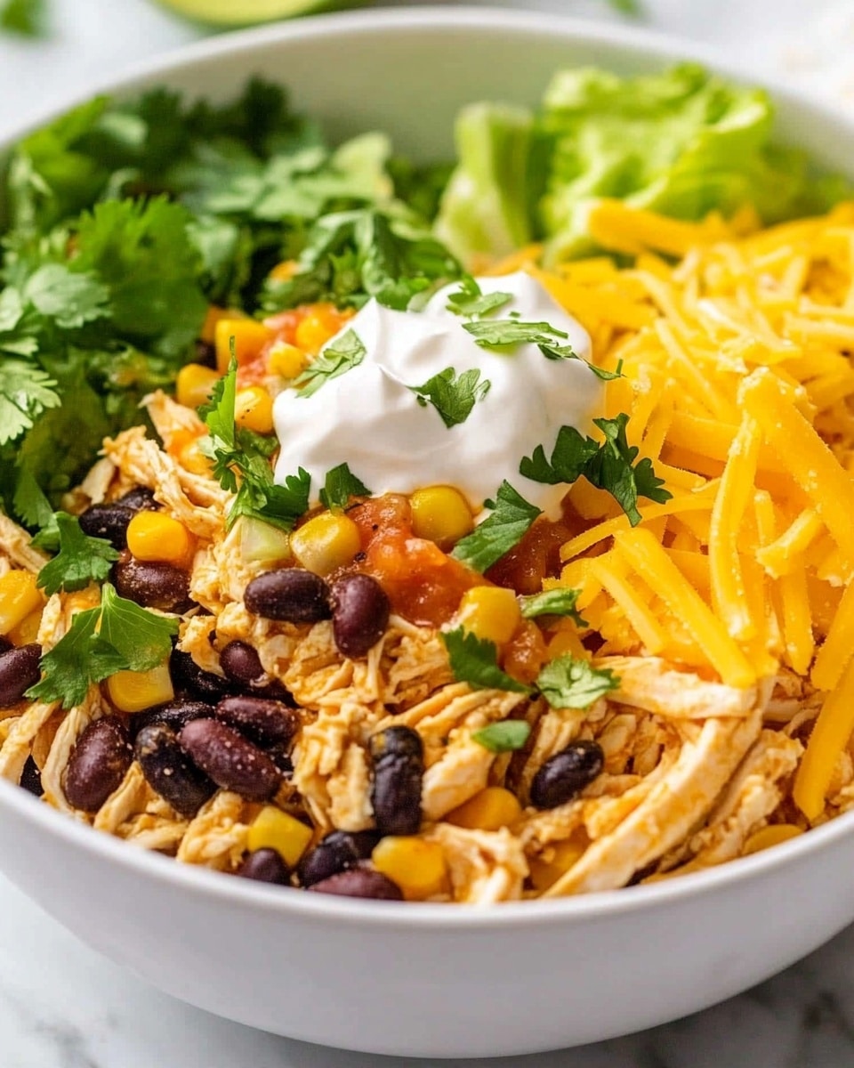 Instant Pot Santa Fe Chicken Burrito Bowls, easy burrito bowl recipes, healthy chicken dinner ideas, weeknight meal prep, flavorful Instant Pot recipes The image shows a white bowl filled with a layered dish starting with a base of shredded light beige chicken mixed with yellow corn kernels and black beans, topped with small bits of red tomato. On top of this mixture is a layer of bright shredded orange cheese scattered unevenly. Fresh green cilantro leaves are sprinkled throughout the dish, adding a touch of freshness. At the edge of the bowl, there is a wedge of green lime resting on the food. The bowl is placed on a white marbled surface with a black and white striped cloth partially visible on the side.