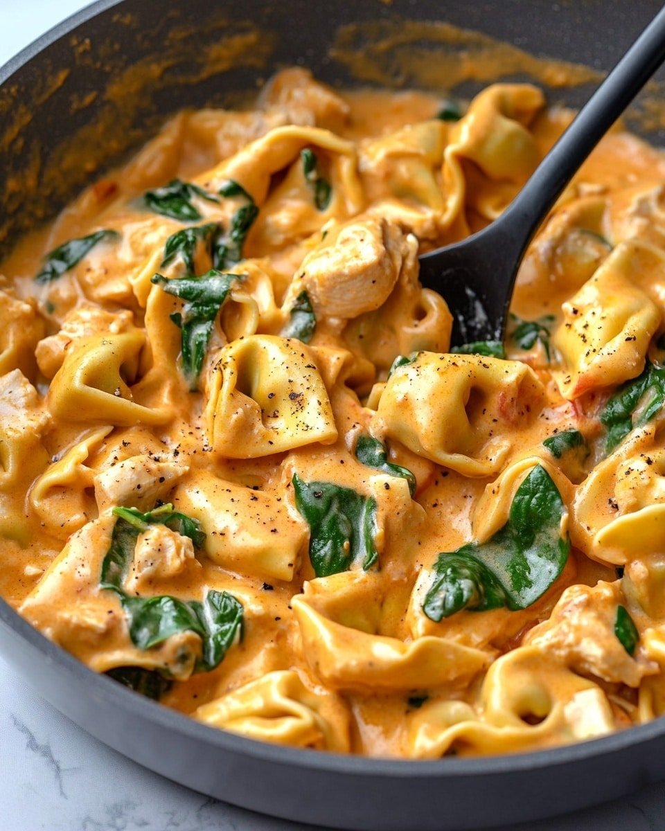 A close-up shot of a creamy pasta dish in a gray pan, showing about three layers mixed together: the first layer is folded tortellini pasta with a smooth, orange creamy sauce; the second layer contains small pieces of cooked chicken with a light browned texture; the third layer has fresh green spinach leaves scattered through the pasta and sauce, adding a pop of color. A black spoon rests in the pan, slightly covered in sauce, and the background is a white marbled texture. photo taken with an iphone --ar 4:5 --v 6.1