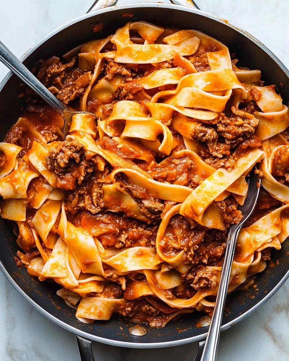 Short Ribs with Pappardelle, Italian short ribs recipe, braised short ribs with pasta, slow-cooked beef pasta dish, easy gourmet short ribs A large pan filled with wide, flat pasta noodles mixed with shredded dark brown meat and a reddish-orange sauce coating the pasta evenly. The pasta sheets appear soft and slightly curled at the edges, layered throughout the pan with chunks of tender meat dispersed evenly in between. Two silver utensils rest on the pasta, one partially lifting some noodles, showing the noodles’ smooth and glossy texture. The pan sits on a white marbled surface.