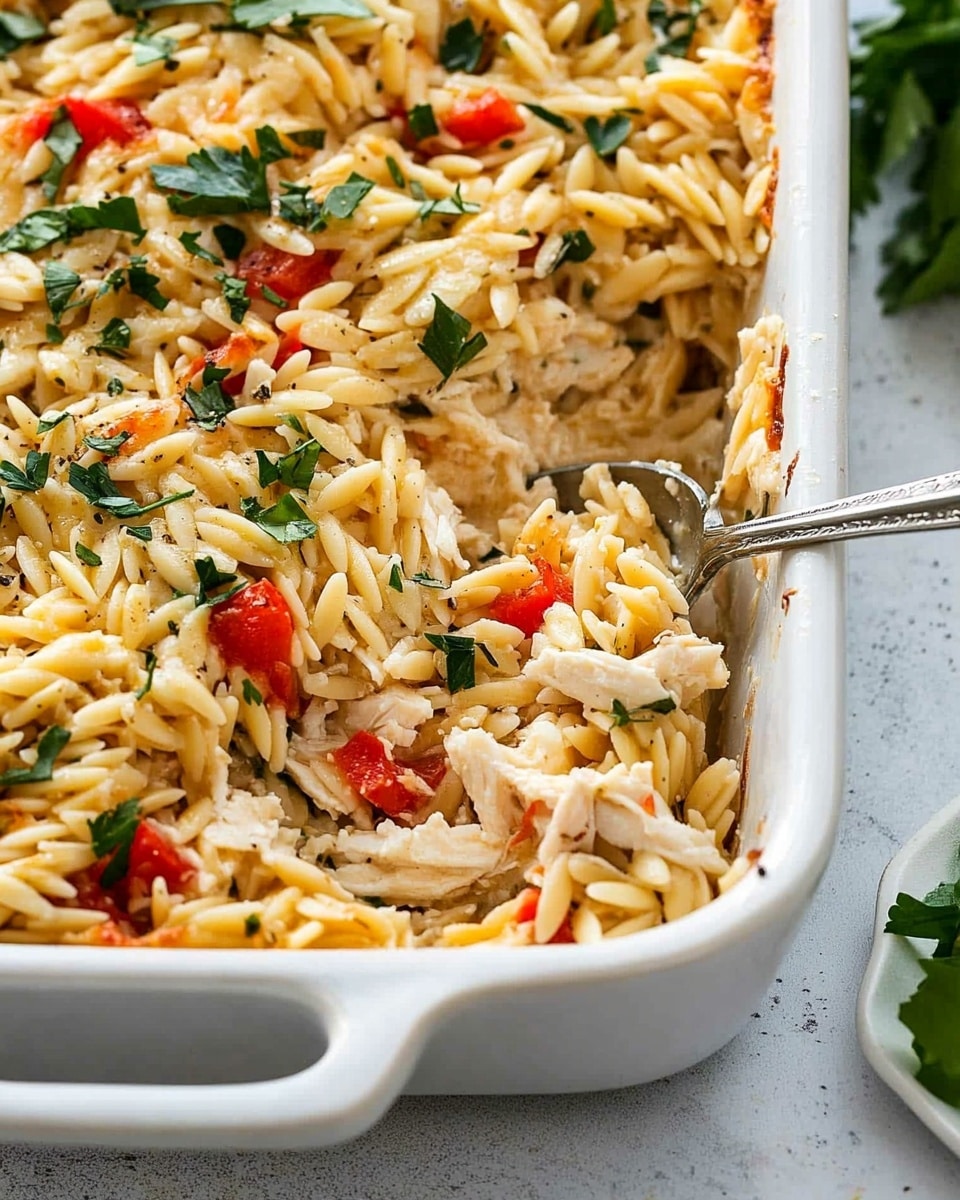 Greek Chicken Casserole with Orzo, Greek Chicken casserole with feta and sun-dried tomatoes, easy Greek chicken bake, Mediterranean chicken pasta bake, flavorful chicken and orzo casserole The image shows a close-up of a white rectangular baking dish filled with a mixed pasta casserole. The dish has one visible thick handle on the near side. The casserole is full of small pasta pieces mixed with shredded light-colored chicken, chopped red bell peppers, leafy green herbs, and small bits of white cheese sprinkled on top. The texture looks creamy with a mix of soft and slightly chunky ingredients. A silver spoon is partly inserted into the casserole on the right side. The dish is placed on a white marbled surface with a small white dish holding some green leaves partially visible on the right edge.