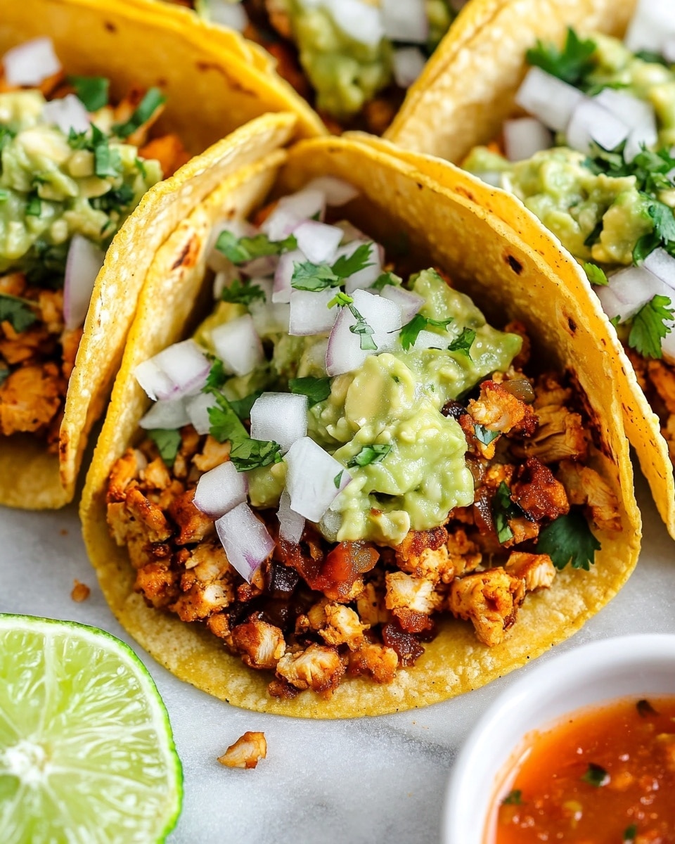 Chicken Street Tacos with Onion Salsa, Easy Chicken Street Tacos, Street-Style Chicken Tacos Recipe, Fresh Onion Salsa for Tacos, Quick Chicken Tacos Three tacos are shown close together on a white marbled surface, each with a soft yellow corn tortilla as the base, lightly toasted with slight browning spots. Inside the tortillas are layers starting with small pieces of cooked, seasoned meat that is reddish-brown. On top of the meat, there is a mix of white chopped onions and green cilantro, giving a fresh and colorful look. A dollop of green guacamole sits on one side of each taco, adding a creamy texture. In the background, a small round white bowl holds a reddish sauce, slightly blurred to keep focus on the tacos. The photo was taken with an iphone