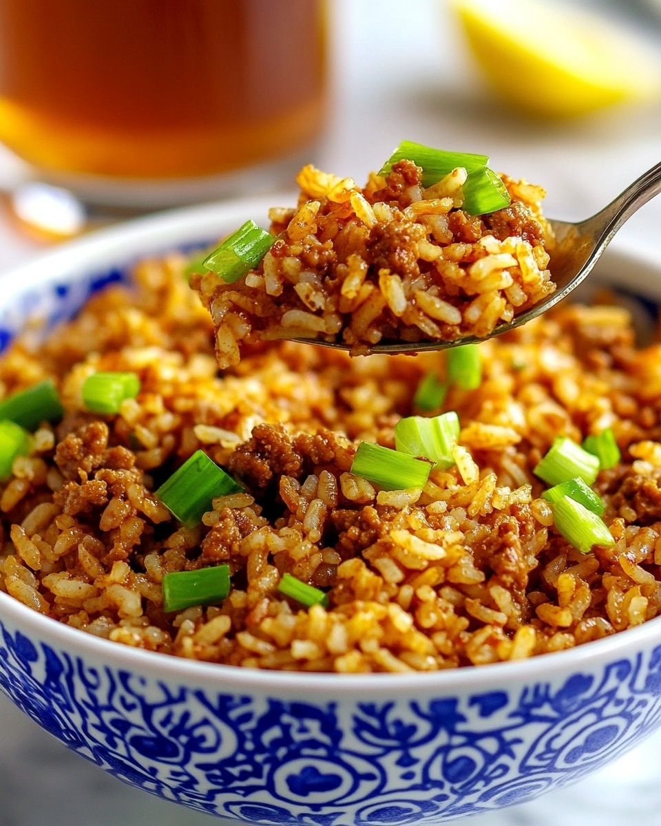 Easy Cajun Sausage Rice, Cajun Sausage Rice, Dirty Rice, Cajun rice recipe, Sausage and Rice dinner A close-up of a white bowl with blue patterns filled with cooked rice mixed with browned ground meat and green chopped scallions scattered on top. A spoon lifted by a woman's hand holds some of the rice and meat mixture above the bowl. The rice looks fluffy with a reddish-brown color from spices, and the bowl is placed on a white marbled surface. In the background, there is a blurred glass of iced tea with a lemon wedge.