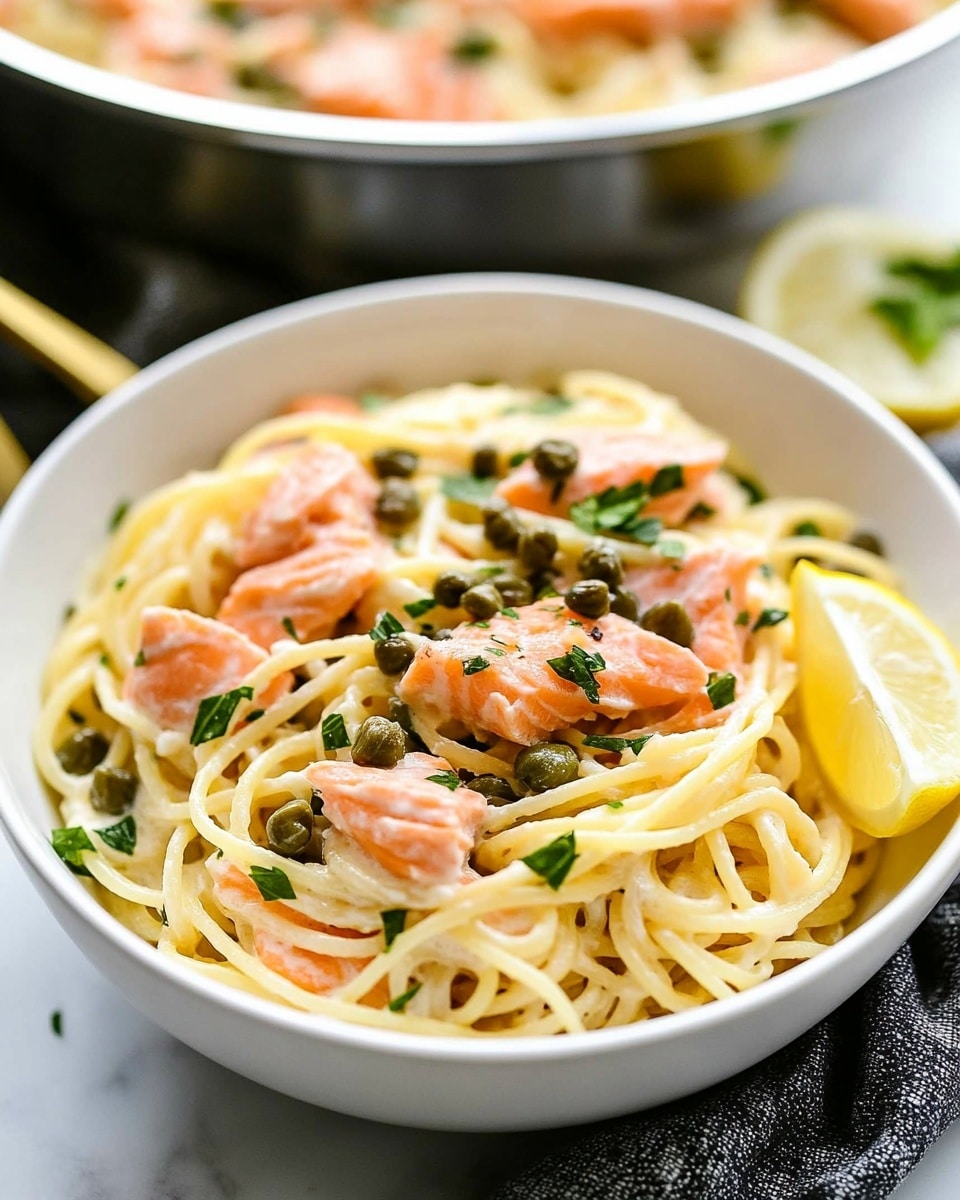A white bowl filled with creamy spaghetti noodles forms the base layer, topped with pink flaky pieces of cooked salmon scattered evenly. Small green capers and chopped fresh green herbs sit on top, adding texture and color contrast. On one side of the bowl, a bright yellow lemon wedge leans against the pasta. In the blurred background, a white pan filled with similar creamy pasta and salmon rests on a dark gray textured cloth, with additional lemon wedges nearby on the white marbled surface. A gold fork partly visible beside the bowl adds a touch of elegance. Photo taken with an iphone --ar 4:5 --v 6.1