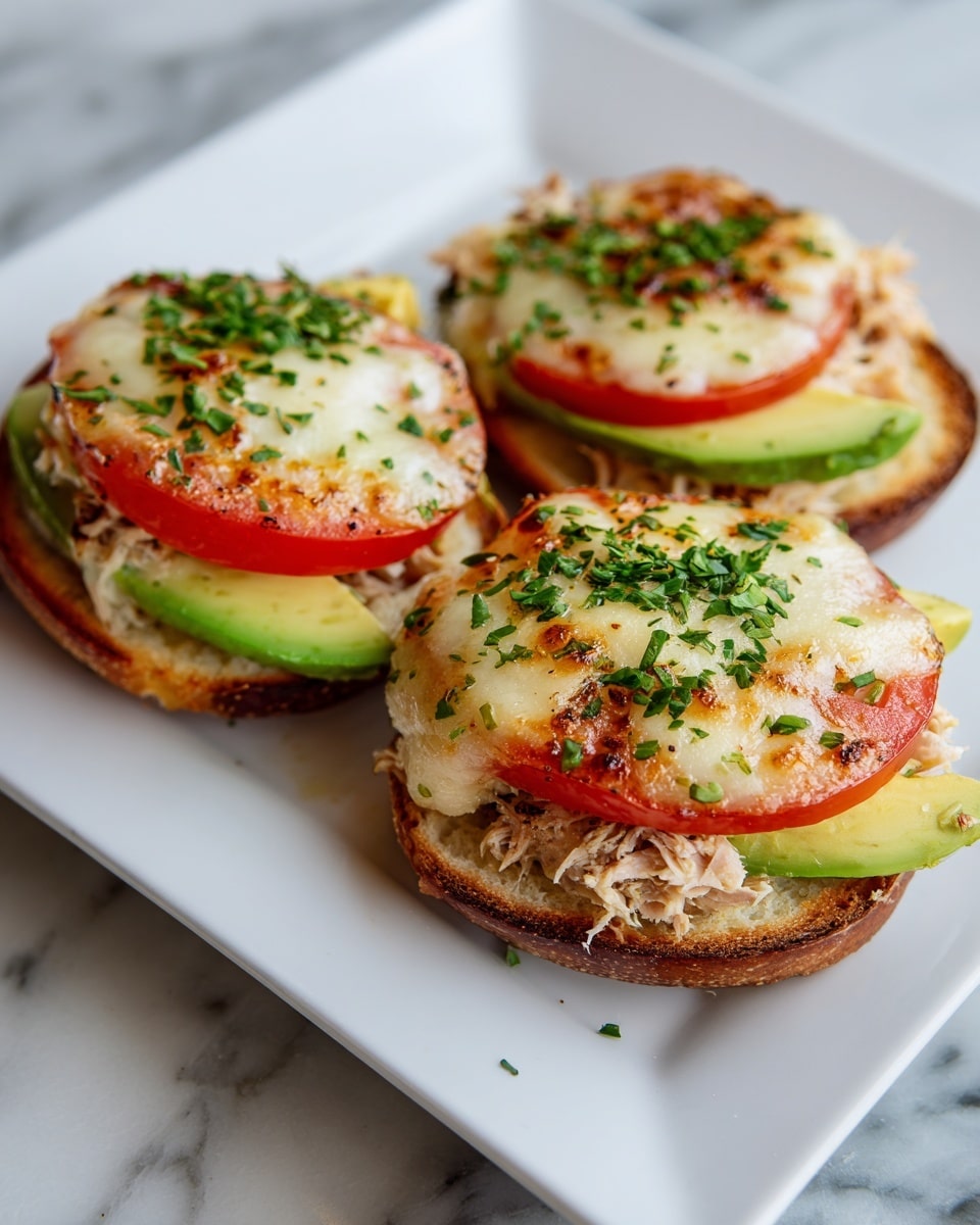 The image shows four open-faced sandwiches on a white plate, each with three visible layers. The bottom layer is a toasted round bread with a golden-brown color. Above that is a layer of fresh tomato slices in bright red and avocado slices in light green with a smooth texture, arranged side by side. The top layer is a creamy, melted cheese mixture with a light golden color and some herbs sprinkled on top, adding small green spots. The plate sits on a white marbled surface with a blurred lemon slice on a wooden board in the background. Photo taken with an iphone --ar 4:5 --v 7