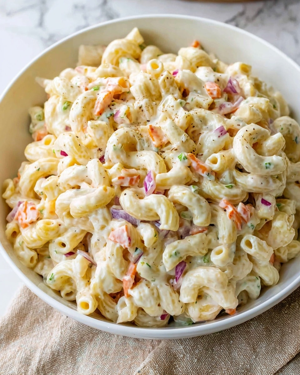A white bowl filled with creamy macaroni salad, showing three main layers: the base layer of curved elbow macaroni in pale yellow, mixed with small chunks of red onion and pieces of orange carrot, scattered green herbs add color throughout, all coated evenly in a smooth, light cream sauce giving a glossy texture. The background is a white marbled surface with a beige cloth visible on the side. Photo taken with an iphone --ar 4:5 --v 7