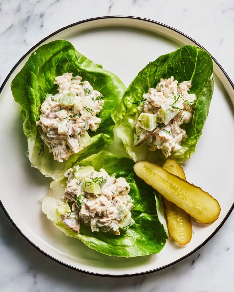 A white plate with a thin black rim holds three bright green lettuce leaves arranged in a triangle shape. On each leaf, there is a creamy white salad with small chunks of light pink and green ingredients mixed in, giving the salad a textured, chunky look. To the upper right side of the plate, there are two thin, pale yellow pickle slices with visible crinkle marks. The plate sits on a white marbled surface. photo taken with an iphone --ar 4:5 --v 7