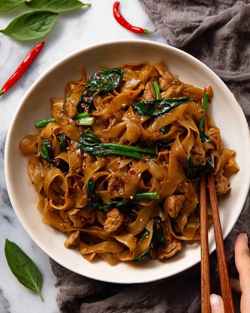 A white bowl filled with three main layers: wide, flat brown noodles coated in a glossy sauce as the base layer; tender pieces of light brown cooked meat scattered throughout the noodles as the second layer; and green vegetable stalks and leaves, including spring onions and basil, mixed in on top, giving a fresh contrast to the dish. The noodles and meat have a slight shine and small specks of red chili pepper flakes are visible. A pair of wooden chopsticks rests on the right side of the bowl, held by a woman's hand. The bowl sits on a white marbled surface with a grey cloth and some red chili peppers and green leaves in the background. Photo taken with an iphone --ar 4:5 --v 7
