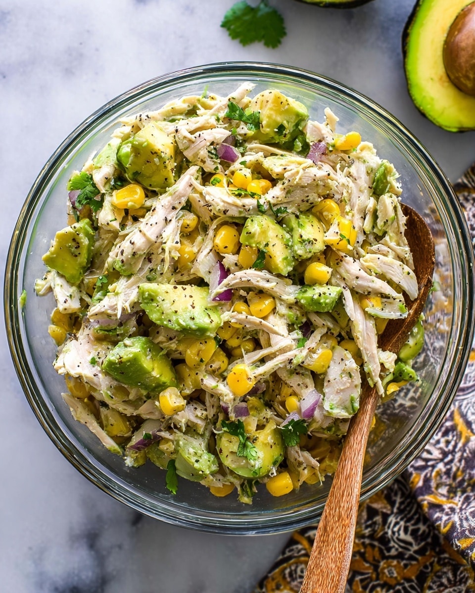 A clear glass bowl filled with a mixed salad showing three main layers: shredded white chicken at the base, vibrant green avocado chunks spread evenly throughout, and small yellow corn kernels dotted all around. Mixed in are small pieces of purple onion and fresh green cilantro leaves. The textures vary from soft avocado to tender chicken and crisp corn, all lightly speckled with black pepper. A wooden spoon rests on the right edge of the bowl. The bowl sits on a white marbled texture with part of an avocado visible in the upper right corner and a patterned cloth in the bottom right corner. Photo taken with an iphone --ar 4:5 --v 7