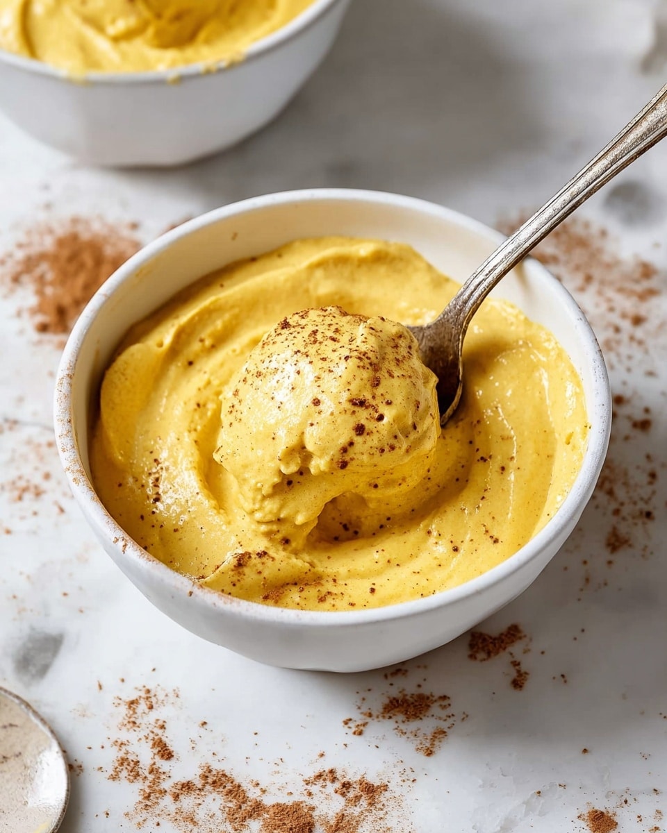 A white bowl filled with a thick, creamy, bright orange-yellow mousse that has a smooth, slightly glossy surface with small air bubbles. A vintage silver spoon scoops into the mousse, lifting a large, rounded dollop showing a soft, dense texture. The bowl sits on a white marbled surface with some sprinkling of brown powder scattered nearby. Another bowl with the same mousse, smooth on top, is partially visible at the top right corner of the image. A beige cloth is placed on the bottom right side, adding a soft fabric contrast. photo taken with an iphone --ar 4:5 --v 7