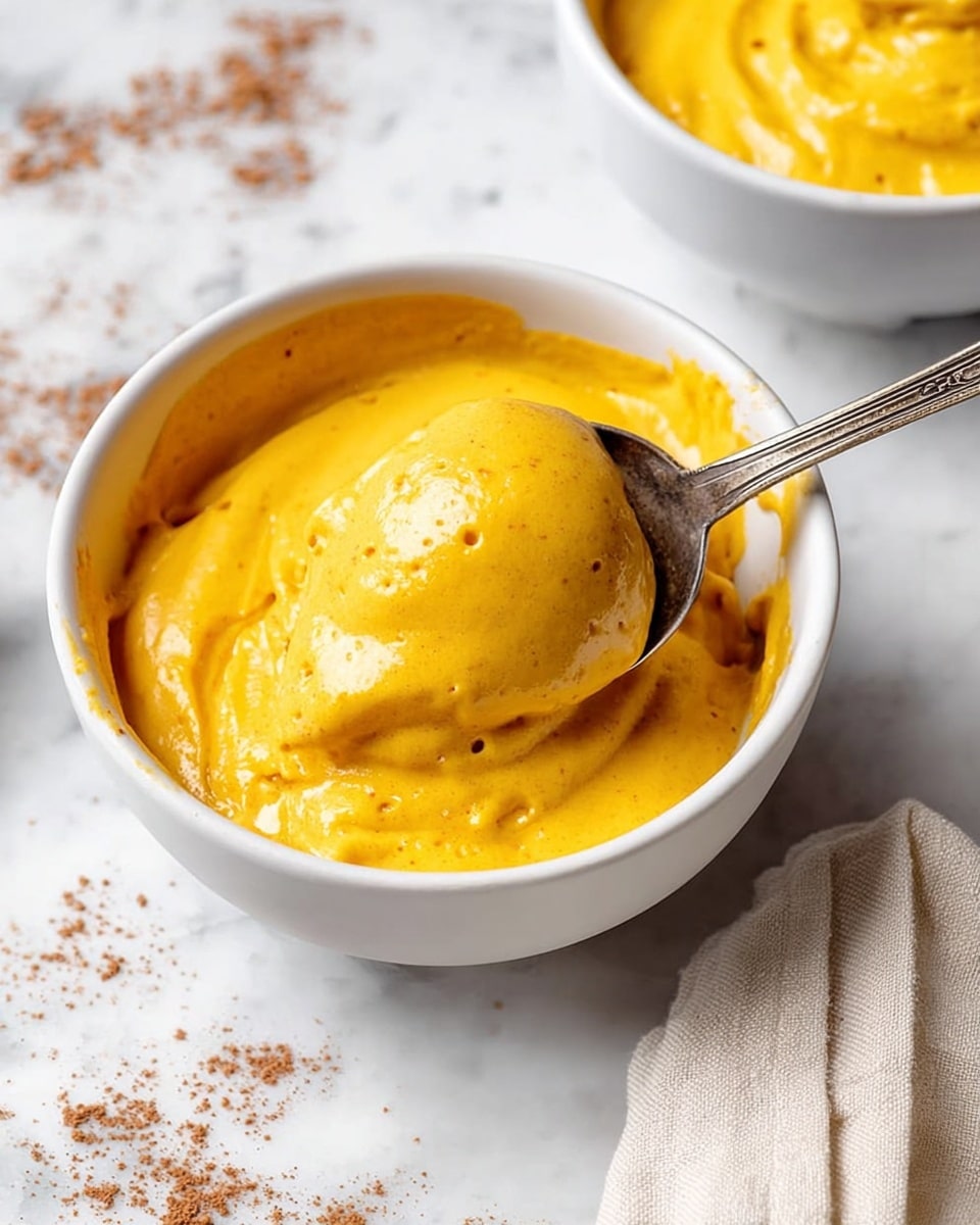 A white bowl filled with one layer of thick, smooth, and creamy bright yellow sauce with a slightly shiny surface, sprinkled lightly with dark brown powder on top. A silver spoon rests inside the bowl, scooping some of the sauce, creating soft folds and texture. The bowl sits on a white marbled surface with some scattered brown powder around it. Photo taken with an iphone --ar 4:5 --v 7