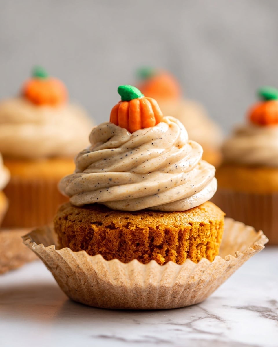 A close-up of one orange cupcake sitting on a white marbled surface, with its light brown paper liner partly unwrapped around the bottom. The cupcake has two layers: the bottom layer is the moist textured orange cake, and the top layer is thick swirled beige frosting with dark specks, shaped in a tall spiral. On top of the frosting, there is a small candy shaped like an orange pumpkin with a green stem. In the blurry background, other cupcakes with the same frosting are visible. photo taken with an iphone --ar 4:5 --v 7
