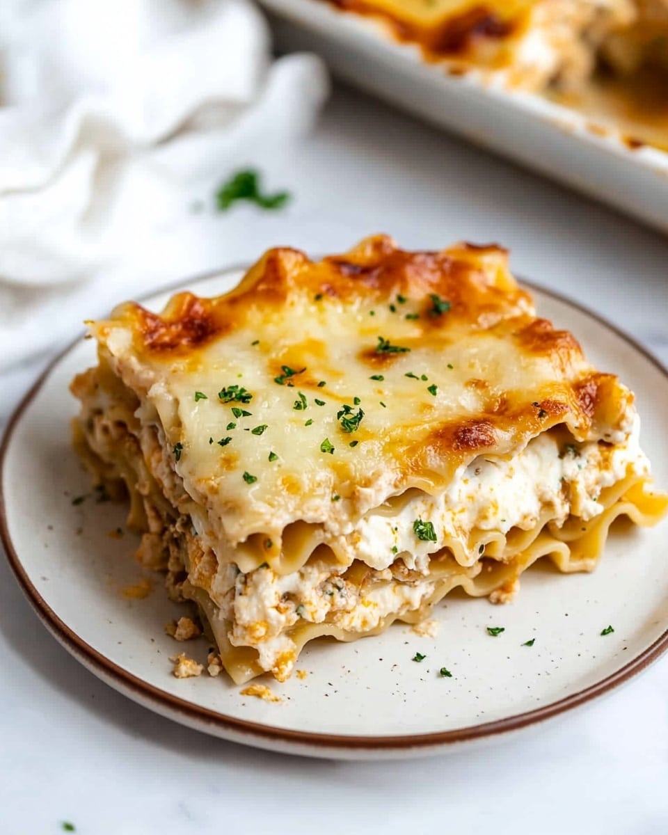 A slice of lasagna is placed on a white plate with a thin brown edge, resting on a white marbled surface. The lasagna has three visible layers of wavy-edged pasta, each separated by creamy white sauce. The top layer is golden brown with melted cheese that has a slightly crispy texture and is sprinkled with finely chopped green herbs and grated white cheese. The edges of the pasta are soft and slightly curled. In the background, a second plate with more lasagna and a white textured cloth are softly blurred. Photo taken with an iphone --ar 4:5 --v 6.1