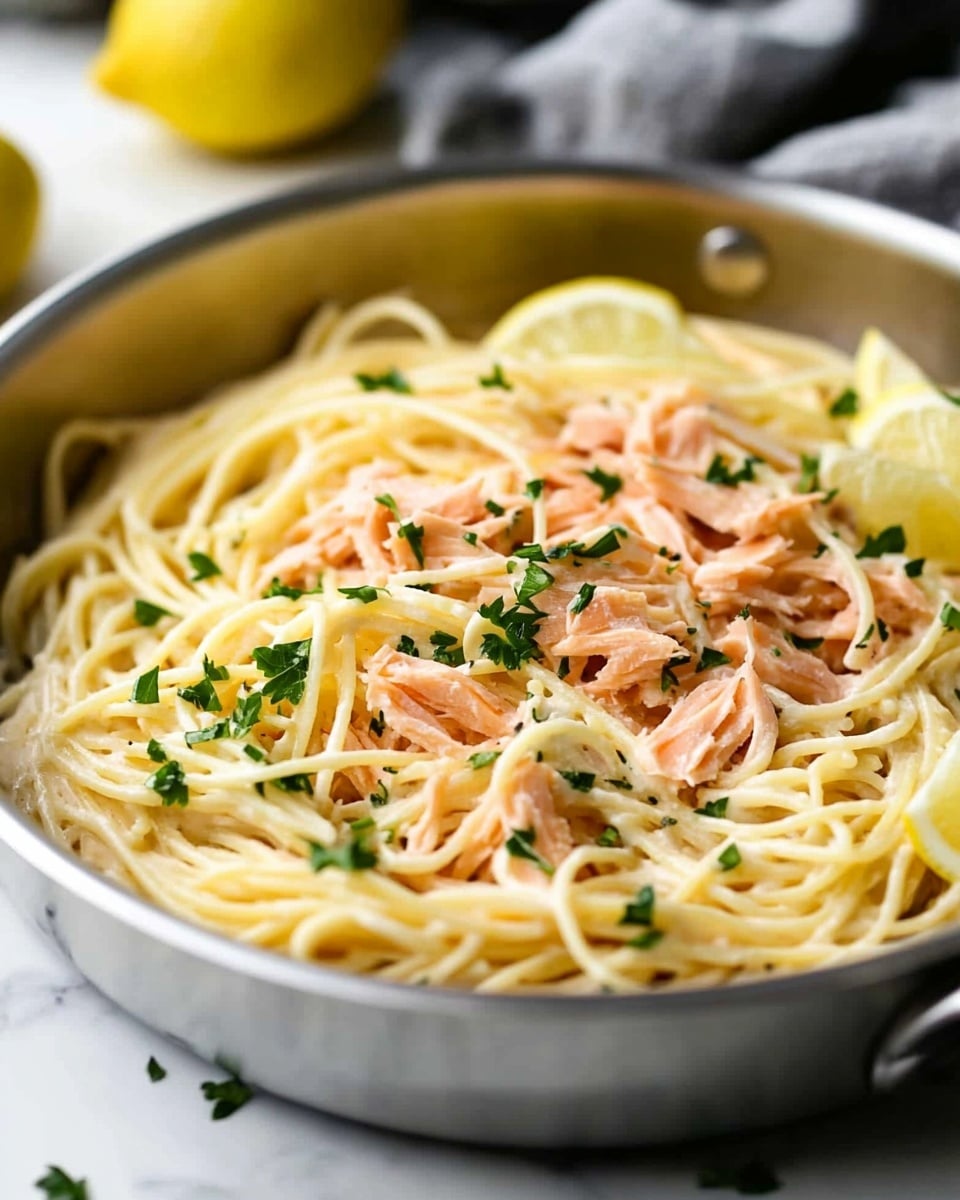 A shiny stainless steel pan on a white marbled surface holds a creamy pasta dish. The base layer is a tangle of light yellow spaghetti noodles, covered with shredded pieces of pink salmon. On top, small green leaves of chopped parsley are scattered, adding a splash of color. On the side of the pan, two thin lemon wedges lean against the noodles. The background shows a blurred yellow lemon and a gray cloth. Photo taken with an iphone --ar 4:5 --v 6.1