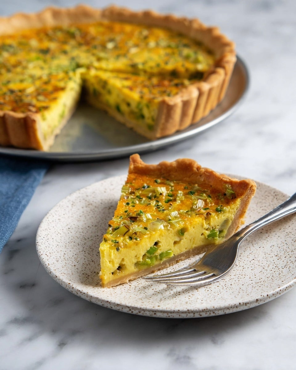 A slice of yellow quiche with green vegetable pieces, possibly green onions or leeks, sits on a white textured ceramic plate with a silver fork placed beside it. The quiche slice has a thick, golden brown crust and a smooth, firm filling dotted with chopped green vegetables and herbs. In the background, the whole quiche, showing roughly six slices with one missing, rests on a round metal tray against a white marbled surface. The quiche’s filling and crust colors are warm, contrasting nicely with the plate and surface. photo taken with an iphone --ar 4:5 --v 7