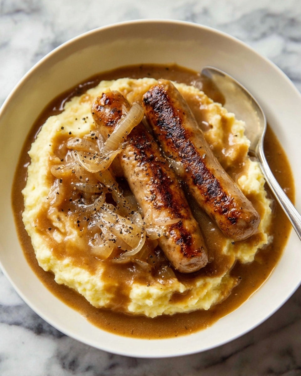 A white bowl holds a bed of creamy, pale yellow mashed potatoes as the base layer, topped with two browned sausages placed diagonally across each other. Over the sausages and mashed potatoes, there is a thick, rich brown gravy with visible cooked onions spread evenly, with a sprinkle of black pepper on top. A silver spoon rests inside the bowl on the right side. The dish is set on a white marbled surface. Photo taken with an iphone --ar 4:5 --v 7