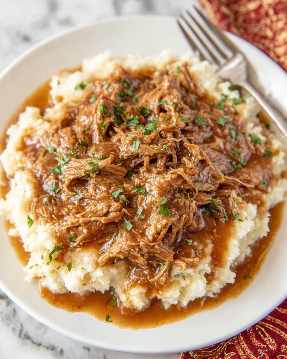 A white plate filled with two layers: the bottom layer is creamy, light beige mashed potatoes with a soft, smooth texture, while the top layer consists of shredded brown meat covered in a thick, slightly shiny brown gravy. The meat is sprinkled with small bits of green herbs, and some of the sauce drips down the sides of the mashed potatoes. The plate sits on a white marbled surface, with a woman's hand holding a silver fork resting nearby. Photo taken with an iphone --ar 4:5 --v 7