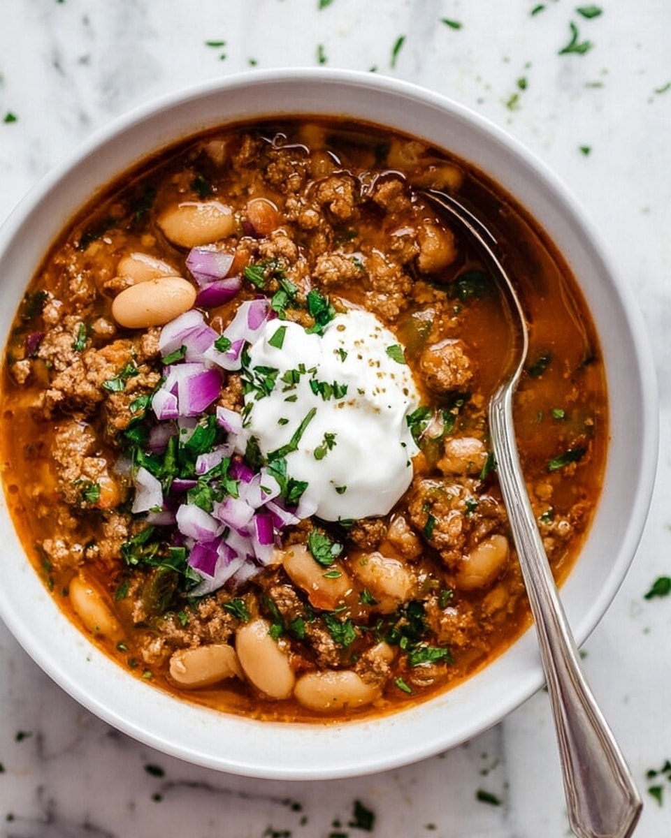 A white bowl filled with thick chili made of cooked ground meat and white beans in a reddish-brown broth, topped with a dollop of white sour cream and sprinkled with chopped purple onions and green herbs. A silver spoon is resting inside the bowl, partially submerged in the chili. The bowl is placed on a white marbled surface with some scattered small green herbs around. photo taken with an iphone --ar 4:5 --v 7