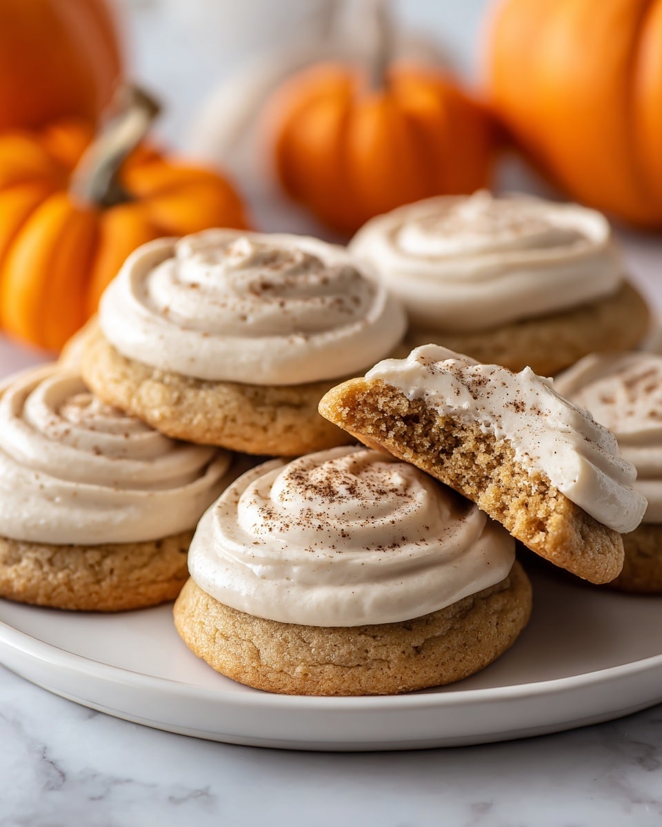 The image shows several round cookies arranged on a wooden board and a white plate with a beige cloth underneath. Each cookie has two layers: a soft golden-brown base and a thick swirl of light beige cream frosting sprinkled with fine brown powder on top. One cookie on the wooden board has a bite taken out, revealing a moist texture inside. There are blurred small orange pumpkins in the background. The whole scene is set on a white marbled surface. photo taken with an iphone --ar 4:5 --v 7