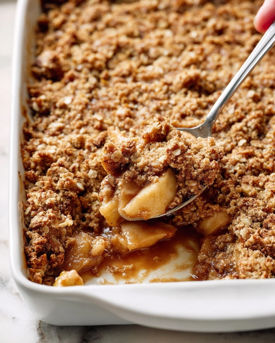 A close-up view of a white baking dish filled with a warm apple crumble, showing two clear layers: the bottom layer is made of soft, cooked apple pieces in a light brown syrupy sauce, while the top layer is a thick, golden-brown oat and cinnamon crumb topping with a crunchy texture. A spoon digs into the crumble, lifting some of the apple filling and crumb mixture, with a woman's hand holding the spoon. The dish rests on a white marbled surface. photo taken with an iphone --ar 4:5 --v 7