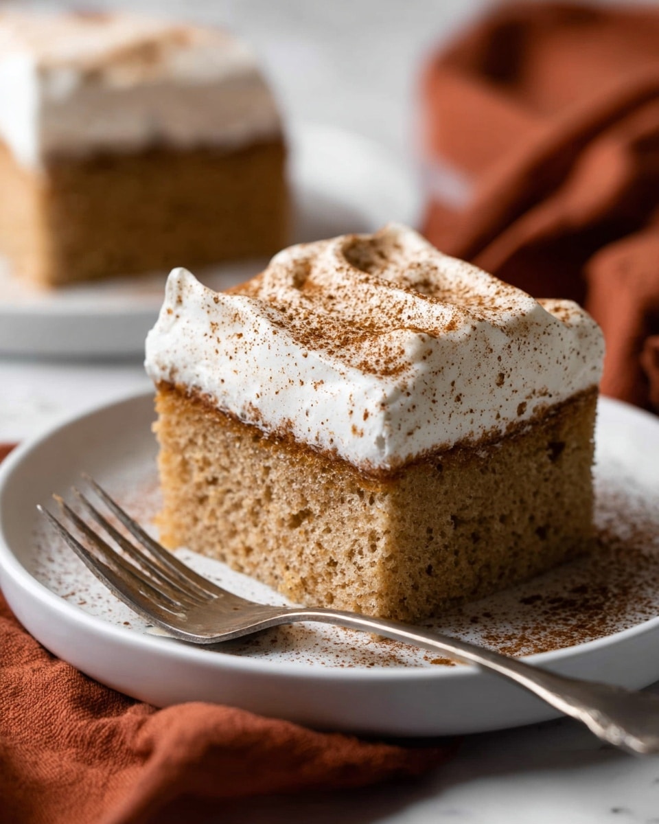 A close-up of a square piece of light brown sponge cake with a moist texture, topped with a thick layer of white cream sprinkled evenly with cinnamon powder. The cake sits on a white plate with a shiny silver fork resting beside it, the plate placed on another white plate. The background shows another similar slice of cake blurred out, all on a white marbled surface with a soft focus on the close cake. In the foreground, a rust-colored cloth is partially visible, adding a warm touch to the scene. Photo taken with an iphone --ar 4:5 --v 7
