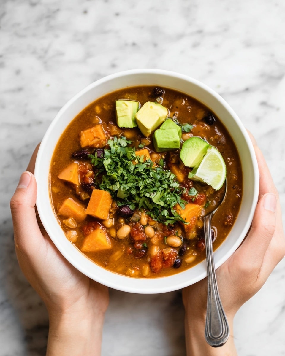 A white bowl filled with thick soup showing chunks of orange sweet potato, white and black beans, and bits of tomato in a brown broth. On top, there are small green cubes of avocado arranged in a line next to finely chopped green cilantro. Two lime wedges with a fresh green color are placed on one side of the bowl. A silver spoon is partially visible inside the bowl on the right side. The bowl is held by two woman's hands above a white marbled surface. photo taken with an iphone --ar 4:5 --v 7