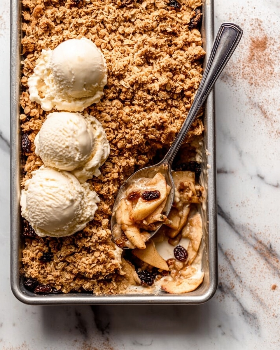 A rectangular metal baking tray filled with a dessert that has three main layers: the bottom layer of soft apple slices, the middle layer with dark raisins, and the top layer of golden-brown oat crumble covering the whole dish. On the left side of the tray, there are two big scoops of pale vanilla ice cream slightly melting on top of the oat crumble. A silver spoon is placed in the right part of the tray, scooping some of the oat crumble with apple slices and raisins. The tray is placed on a white marbled surface with a few small spots of cinnamon powder scattered around photo taken with an iphone --ar 4:5 --v 7