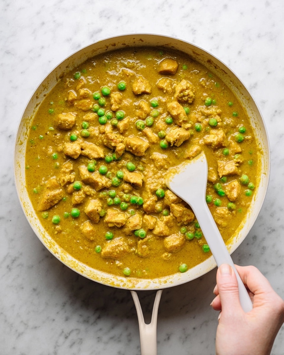 A white pan filled with a thick yellow curry sauce with many small green peas and several uneven light brown chunks of cooked meat or tofu spread evenly throughout. A white spatula is partially submerged in the curry with a woman's hand holding it from the bottom right corner. The pan rests on a white marbled surface, and the curry looks glossy and rich. photo taken with an iphone --ar 4:5 --v 7