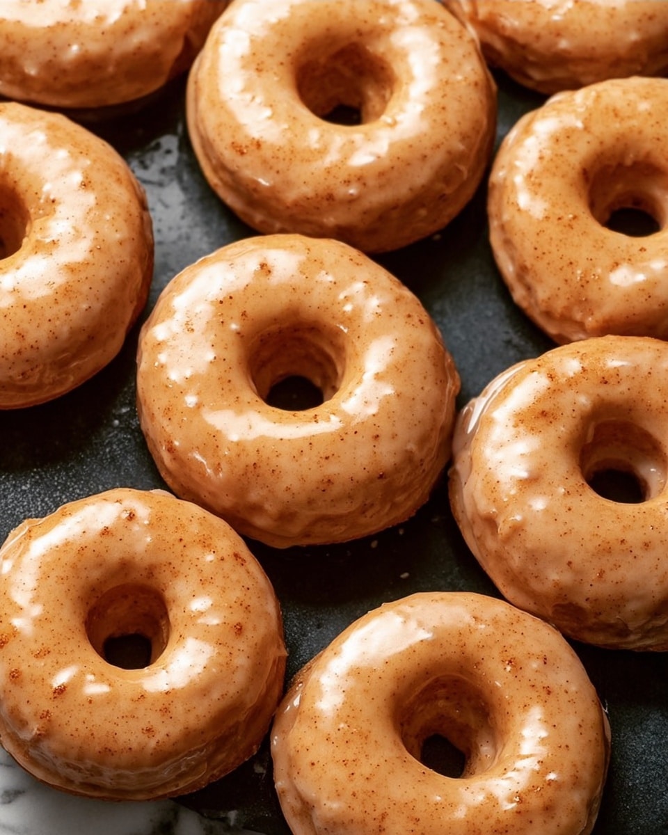 The image shows eight donuts with a shiny, light brown glaze covering their surfaces, giving them a smooth and slightly textured look. Each donut has a thick, even layer of glaze all around, with small darker spots showing through the glaze, adding a bit of texture and color variation. They are arranged closely together on a metal cooling rack, which has thin, shiny silver bars running horizontally and vertically below the donuts. The background surface beneath the rack is a white marbled texture. The lighting is soft and natural, highlighting the glossy finish of the glaze on each donut. Photo taken with an iphone --ar 4:5 --v 7