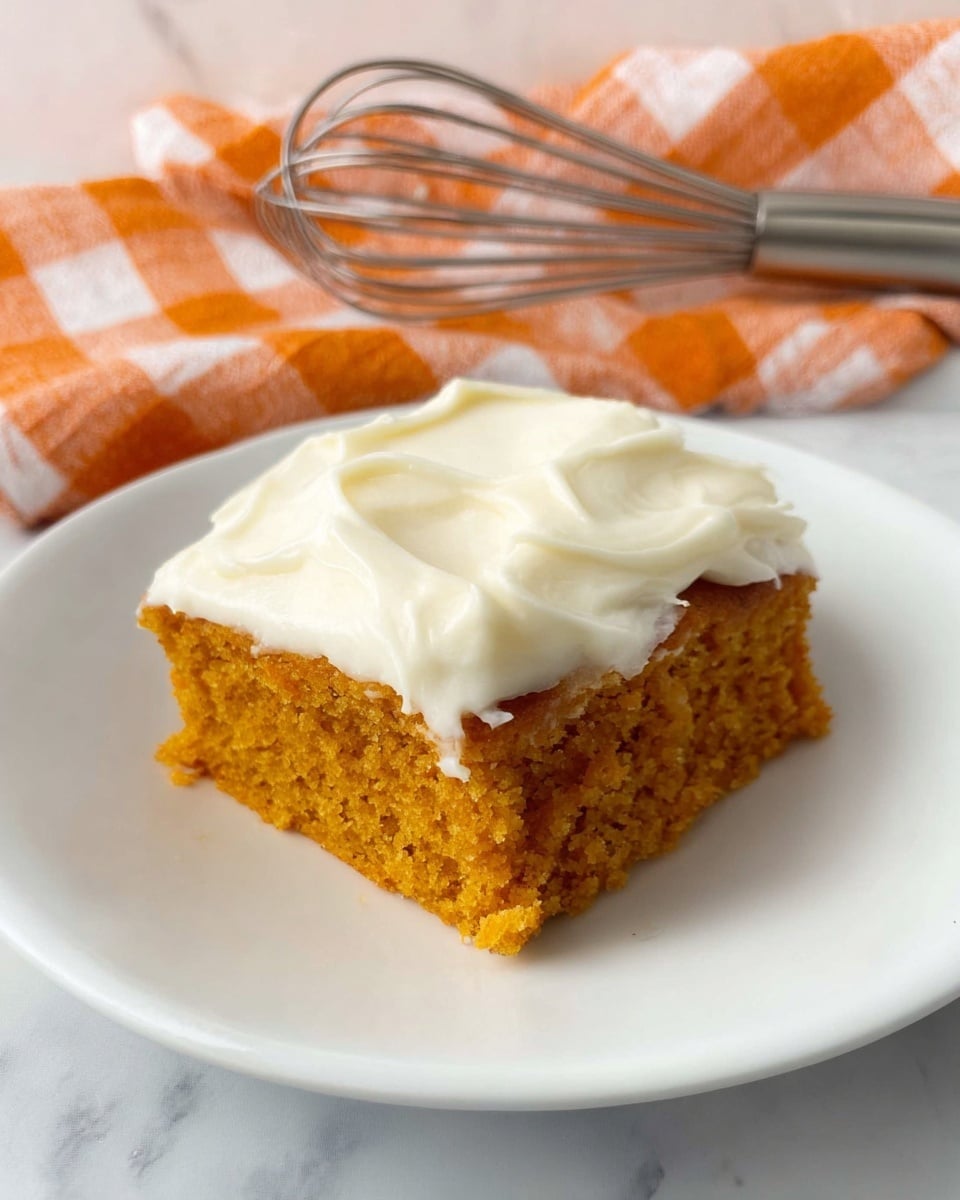 A single square piece of orange cake with a soft, moist texture sits on a white plate. The cake has one thick layer of creamy white frosting spread unevenly on top, with smooth and swirled texture. Behind the plate, there is a shiny metal whisk resting on an orange and white checked cloth. The background surface is white marble. photo taken with an iphone --ar 4:5 --v 7