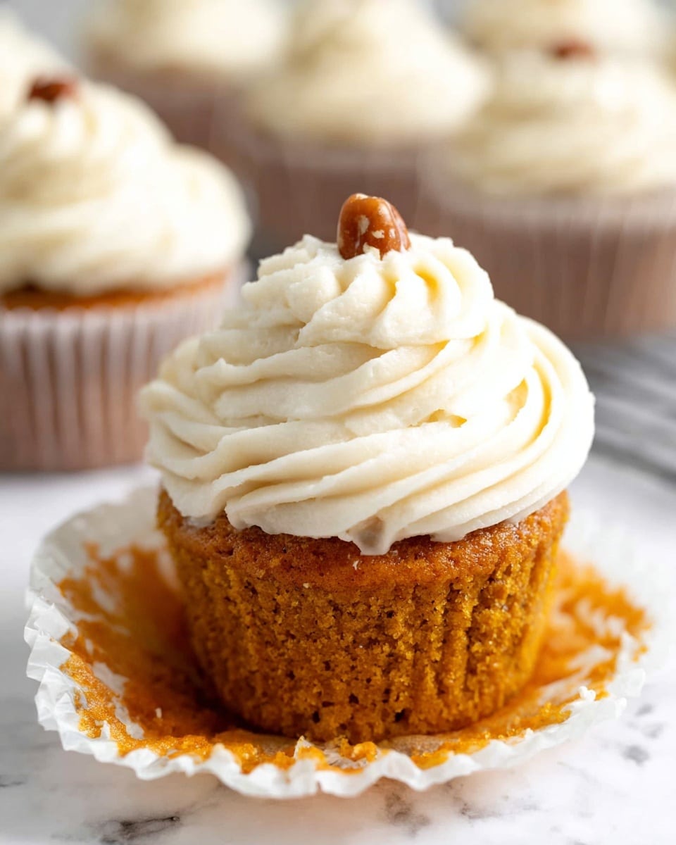 A cupcake with a warm orange base sits at the center, topped with three tall, thick layers of white creamy frosting swirled in a circular pattern. The frosting has small brown specks inside, showing texture and flavor. A single pecan piece rests on top as decoration. The cupcake is shown on a white marbled surface, with blurred similar cupcakes in the background. photo taken with an iphone --ar 4:5 --v 7