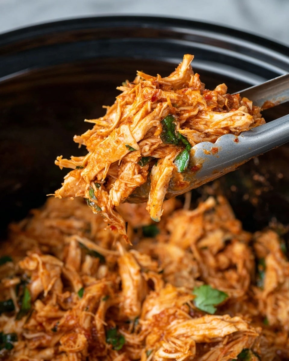 The image shows a close-up of shredded chicken cooked with reddish-orange sauce, giving the meat a moist and tender look. The chicken pieces are small to medium in size, mixed evenly with small bits of herbs, and some green leaves are visible, adding a fresh touch. The chicken is being lifted from a dark pot using silver tongs. The background is blurred black inside the pot, and the scene is set on a white marbled surface. Photo taken with an iphone --ar 4:5 --v 7