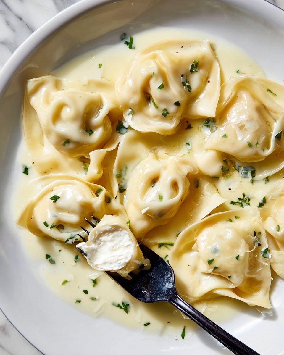 The image shows a white plate with nine pieces of tortellini pasta covered in smooth white cream sauce. The pasta has a pale yellow color with soft, folded shapes creating gentle curves and folds. Green chopped herbs are sprinkled over the pasta and sauce, adding small touches of color. A black fork rests on the right side of the plate, partly under some tortellini. The plate is set on a white marbled surface, and there is a glass of light-colored drink and a blue-gray cloth napkin nearby. photo taken with an iphone --ar 4:5 --v 7