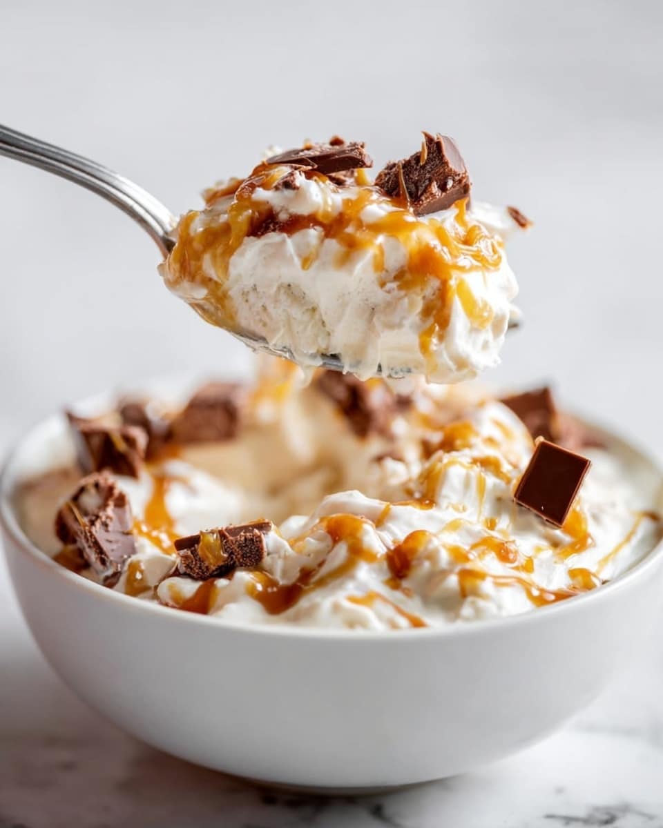A close-up image shows a silver spoon lifting a scoop of creamy white dessert that is swirled with golden caramel sauce and includes small chunks of dark brown chocolate pieces. The dessert is thick and fluffy with a smooth texture, sitting in a white bowl. The bowl is filled mainly with the creamy white base mixed with golden caramel streaks and scattered chocolate chunks. The background is a soft white marbled texture, making the dessert the clear focus. Photo taken with an iphone --ar 4:5 --v 7