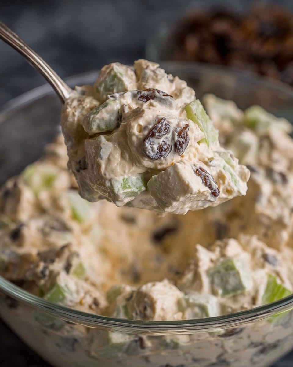 A close-up of a creamy, light beige salad mixture lifted by a spoon above a clear glass bowl filled with the same salad. The salad has a rough texture with visible chunks of light green celery and dark brown raisins mixed evenly throughout. The creamy base looks thick and smooth, holding the chunky ingredients together. The background is softly blurred, showing a hint of a dark surface, while the focus is clearly on the detailed salad on the spoon and in the bowl. photo taken with an iphone --ar 4:5 --v 7