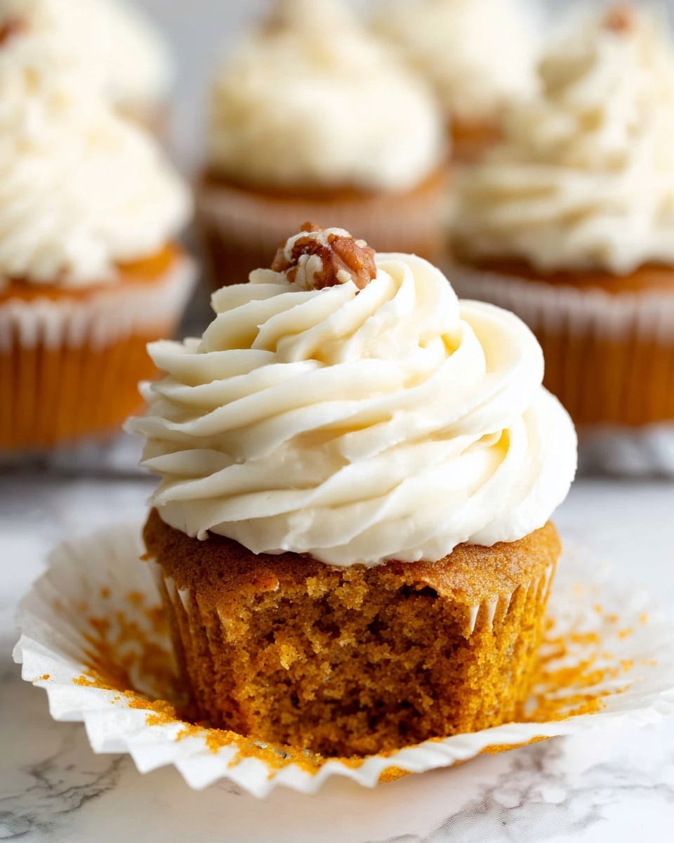 A close-up of a cupcake with an orange-brown base showing a soft and moist texture, topped with a thick swirl of creamy white frosting that has a smooth and slightly textured surface. The frosting is piped in decorative waves and is topped with a single small brown nut. The cupcake is placed on a white paper liner which is slightly peeled down, sitting on a white marbled surface with blurred cupcakes in the background. The photo taken with an iphone --ar 4:5 --v 7