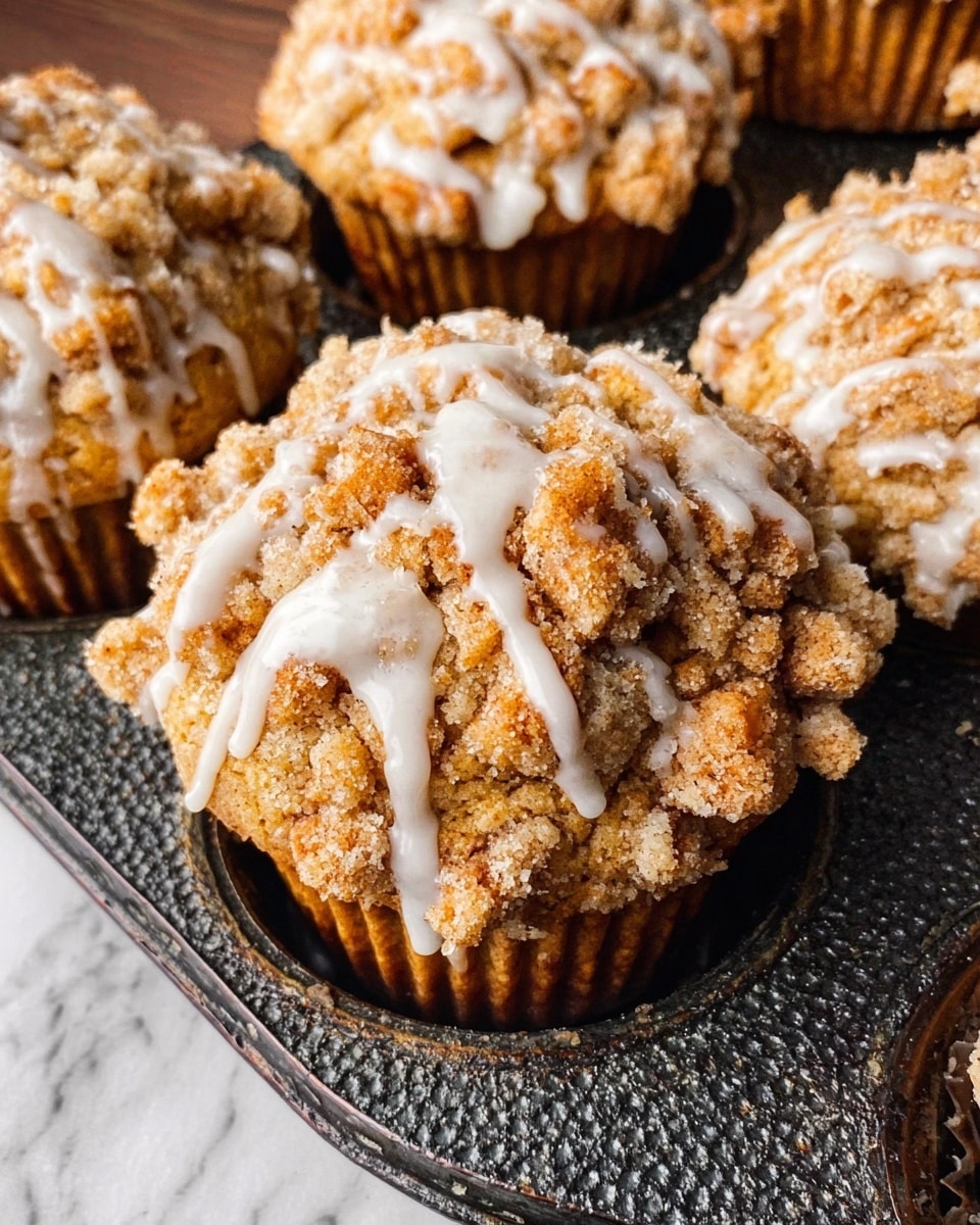 The image shows close-up muffins with a crumbly topping and white glaze drizzled unevenly on top. Each muffin has a golden brown base with a rough texture, topped with chunky crumb pieces in a lighter brown color scattered across the surface. The white glaze creates irregular streaks and droplets over the crumb layer. The muffins are placed on a dark textured muffin tin resting on a white marbled surface. photo taken with an iphone --ar 4:5 --v 7