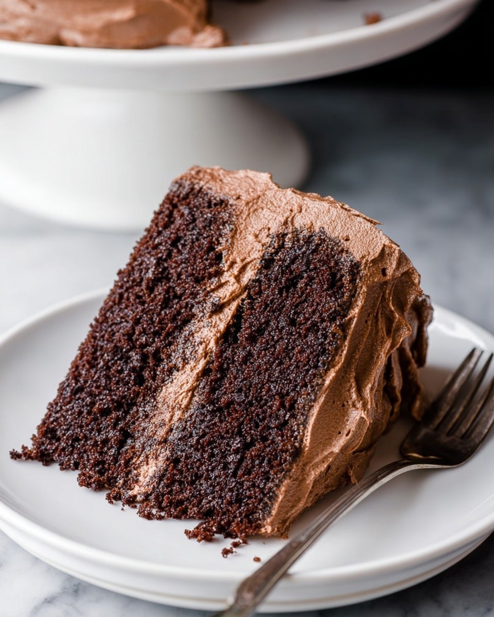 The image shows a two-layer chocolate cake with a thick, smooth layer of chocolate frosting covering the top and sides. The cake layers are dark brown and moist, separated by a generous layer of creamy chocolate frosting. The cake sits on a white cake stand with a beaded edge detail, placed on a white marbled surface. A slice has been removed from the cake, revealing the textured crumb of the cake and the soft, evenly spread frosting between layers. The background is out of focus and dark, making the cake stand out clearly. Photo taken with an iphone --ar 4:5 --v 7
