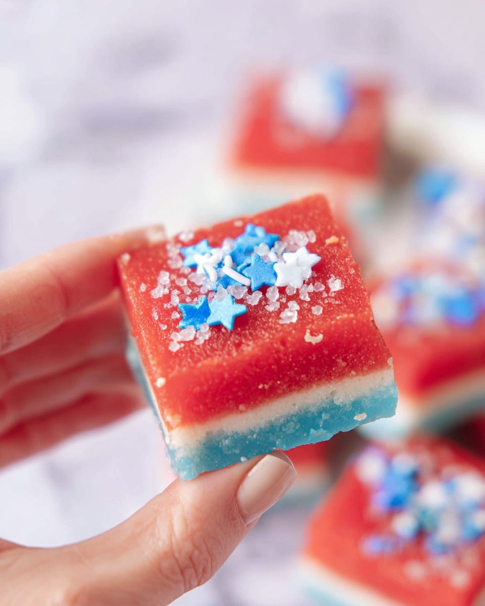A close-up of a small square layered treat held by a woman's hand, showing three clear layers: a bright red top layer adorned with small blue and white star-shaped sprinkles and coarse sugar crystals, a thin white middle layer, and a bottom blue layer visible at the edges; the background has a soft blur with more of the same layered treats on a white marbled surface. Photo taken with an iphone --ar 4:5 --v 7