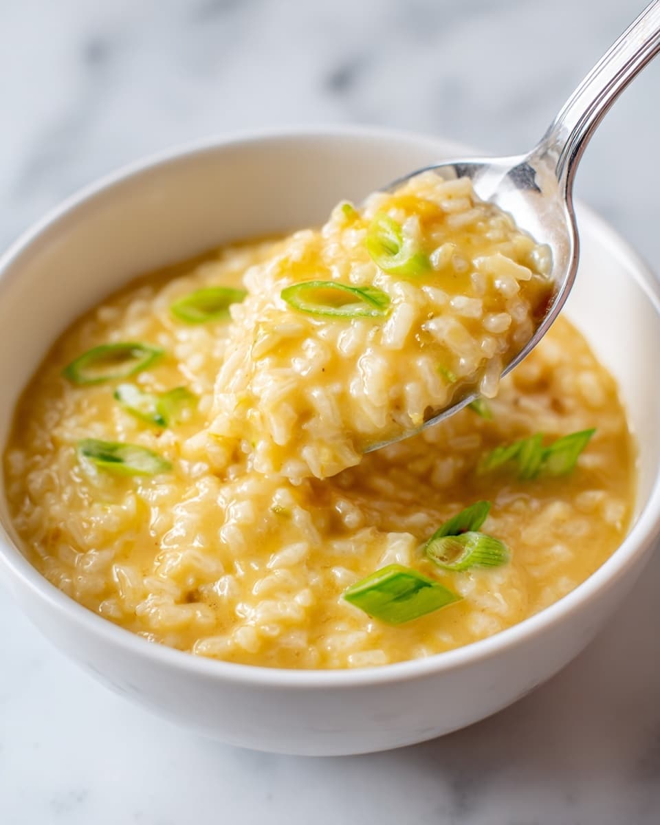 A close-up view of a creamy, soft yellow rice dish served in a white bowl. The dish has a smooth texture with small, tender rice grains mixed in a slightly thick, yellowish sauce. On top, there are small green slices of scallions adding a fresh, bright contrast. A metal spoon lifts a scoop of the rice, showing a mix of the sauce, rice, and scallions in one portion. The background is a white marbled surface. photo taken with an iphone --ar 4:5 --v 7