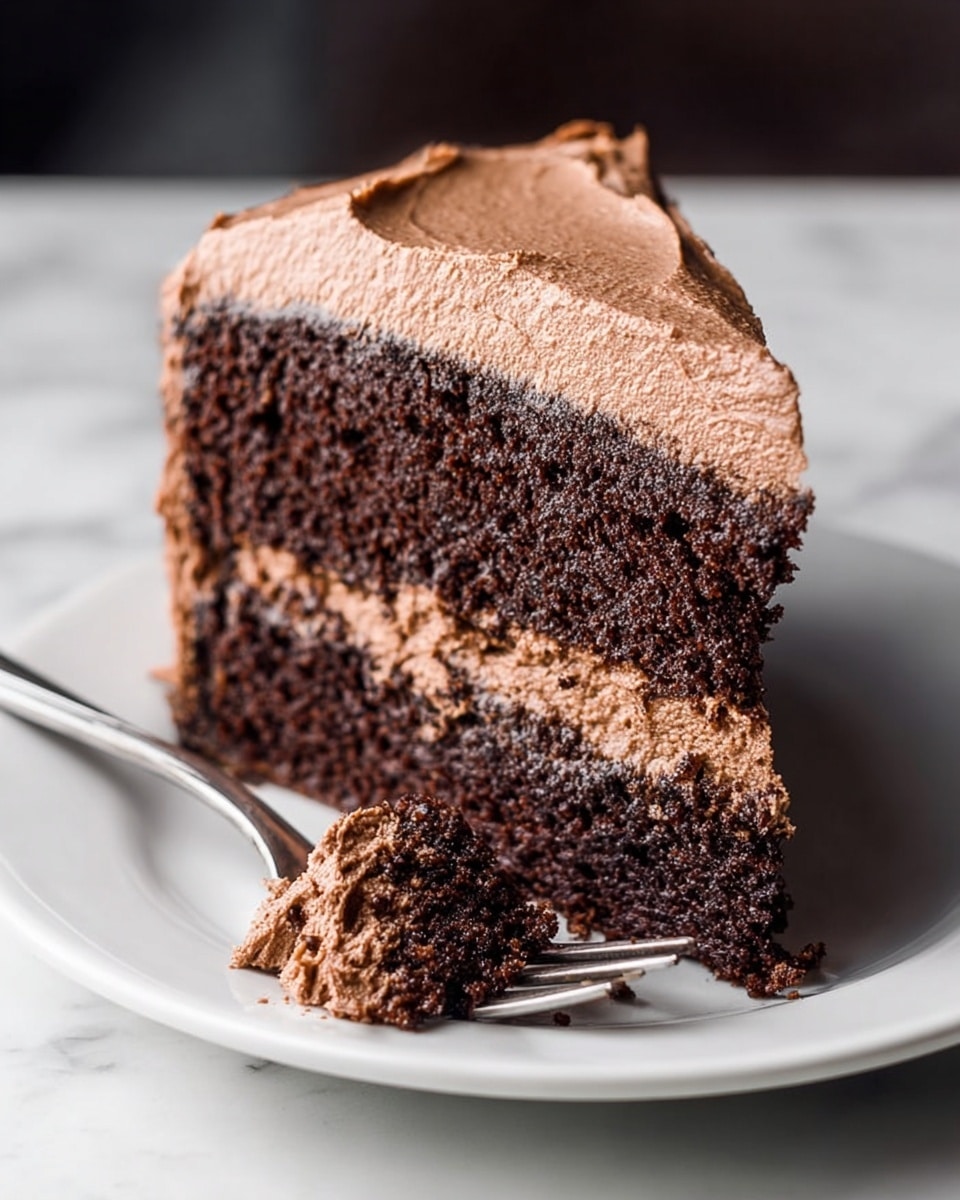 The image shows a close-up of a slice of chocolate cake on a white plate with a white marbled texture background. The cake has two thick dark brown layers that look moist and soft. Between the layers and covering the outside is a thick layer of smooth light brown chocolate frosting with a creamy texture. A small piece of the cake with frosting is on a silver fork that rests on the plate near the slice. The lighting highlights the rich color and texture of the cake and frosting. Photo taken with an iphone --ar 4:5 --v 7
