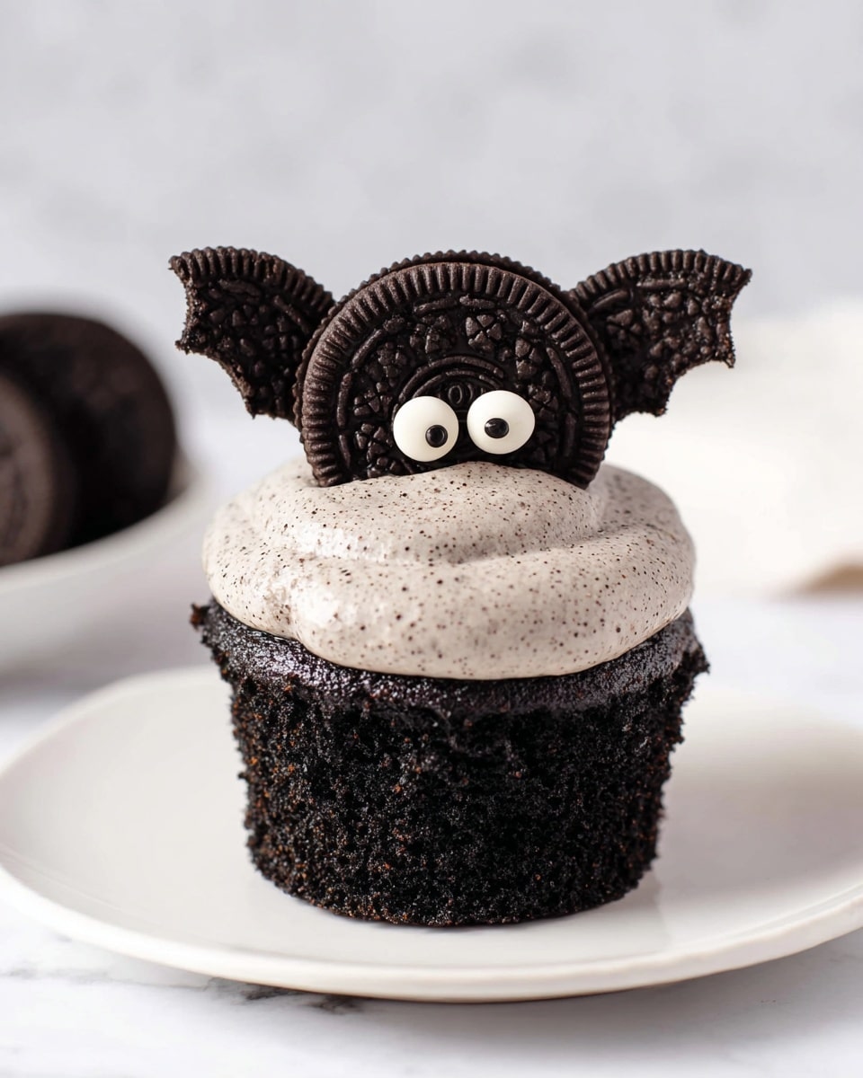 The image shows a black chocolate cupcake with two distinct layers: a dark, moist chocolate cake base and a creamy, light grey frosting with small dark speckles on top. The cupcake is decorated with a whole round chocolate cookie placed vertically at the top center, with two half cookie pieces on each side resembling bat wings. The round cookie features two small white candy eyes with black dots in the middle. The cupcake is on a white plate set on a white marbled surface, with a woman's hand holding a fork nearby in the background. Photo taken with an iphone --ar 4:5 --v 7