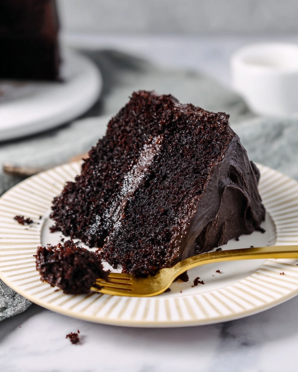 A slice of dark chocolate cake with two thick layers and creamy dark chocolate frosting between and on top, sitting on a white plate with ridged edges. The cake texture looks soft and moist with fine crumbs. A gold fork is holding a small piece of cake near the slice. The plate is placed on a white marbled surface, and the background is softly blurred, showing a white cloth and cake stand. Photo taken with an iphone --ar 4:5 --v 7