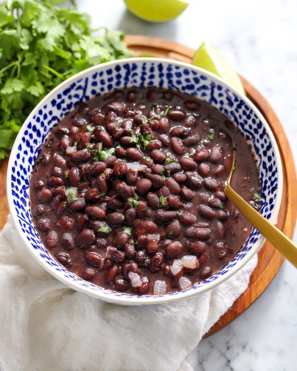 A deep white bowl with blue geometric patterns filled with dark brown cooked black beans in a thick purple-brown sauce. The beans are whole and glossy, mixed with translucent soft small onion pieces. A gold spoon is placed inside the bowl on the right side. The bowl sits on a white cloth over a wooden round board with green cilantro leaves in the top left corner and a lime barely visible at the bottom left. The background is a white marbled texture. photo taken with an iphone --ar 4:5 --v 7