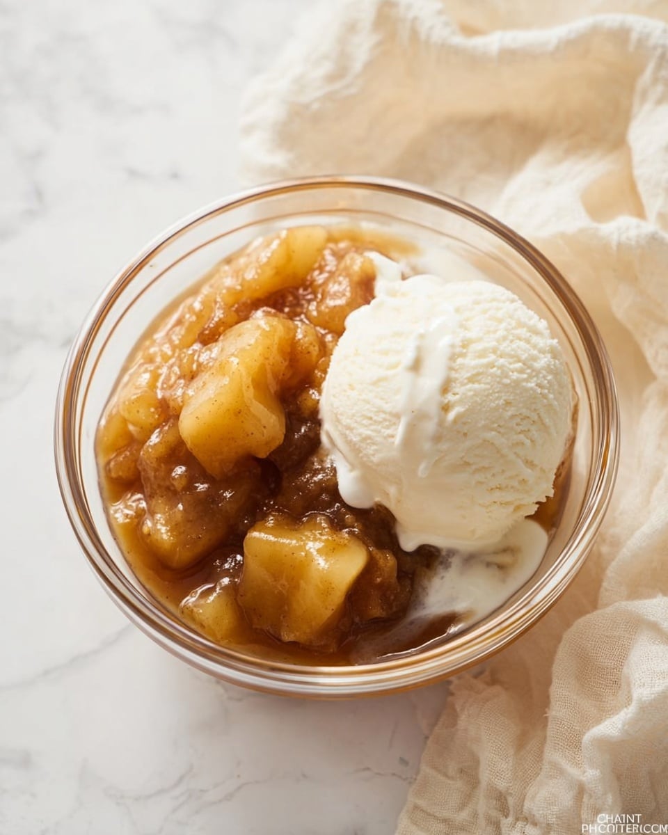 A clear glass bowl on a white marbled surface holds two main layers: on the left, a warm golden-brown apple dessert with soft chunks of apple coated in a shiny syrup, showing a slightly sticky texture, and on the right, a round scoop of smooth, creamy white vanilla ice cream gently melting and blending into the apple layer. The background has a light beige cloth with soft folds. photo taken with an iphone --ar 4:5 --v 7