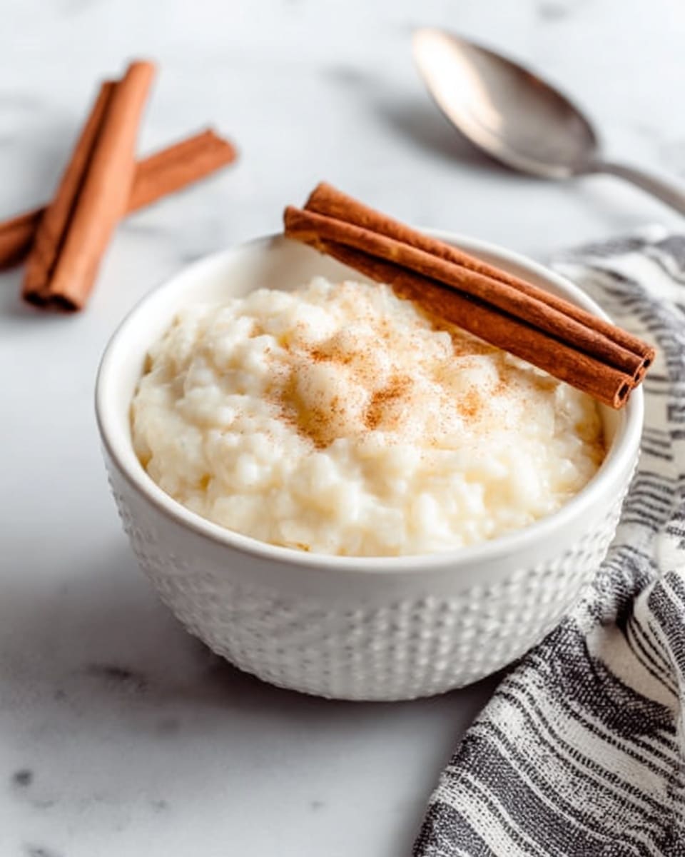 A white bowl filled with creamy, slightly lumpy rice pudding that has a soft off-white color, topped with two long brown cinnamon sticks placed diagonally on one side. The bowl sits on a white marbled surface, with two more cinnamon sticks lying next to it. In the background, there is a white bowl stacked on another white bowl and a partially visible white plate with some rice pudding, all on the same white marbled texture. A silver spoon is placed beside the bowl closer to the bottom left corner. The overall setting is bright and clean with soft lighting. photo taken with an iphone --ar 4:5 --v 7