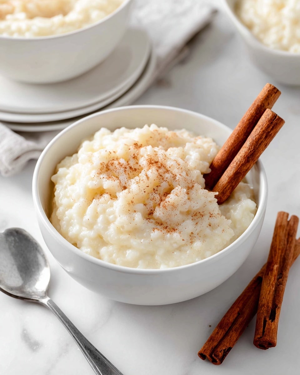 A bowl filled with creamy white rice pudding that has a soft, slightly thick texture, topped with a whole cinnamon stick resting on the right side of the bowl. The bowl is white with a subtle textured pattern on the outside, sitting on a white marbled surface. To the left of the bowl, there is another cinnamon stick lying flat and a silver spoon partially visible behind it. On the right side of the frame, a soft striped cloth in black and white is partially seen. Photo taken with an iphone --ar 4:5 --v 7