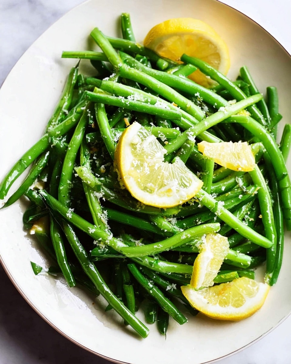 The image shows a white plate filled with bright green green beans arranged in a loose pile. Among the green beans, there are a few lemon wedges with yellow skin and pale yellow inside, adding a pop of color. The green beans have a shiny, slightly wet texture with small grated white bits sprinkled on top. The plate sits on a white marbled surface. photo taken with an iphone --ar 4:5 --v 7