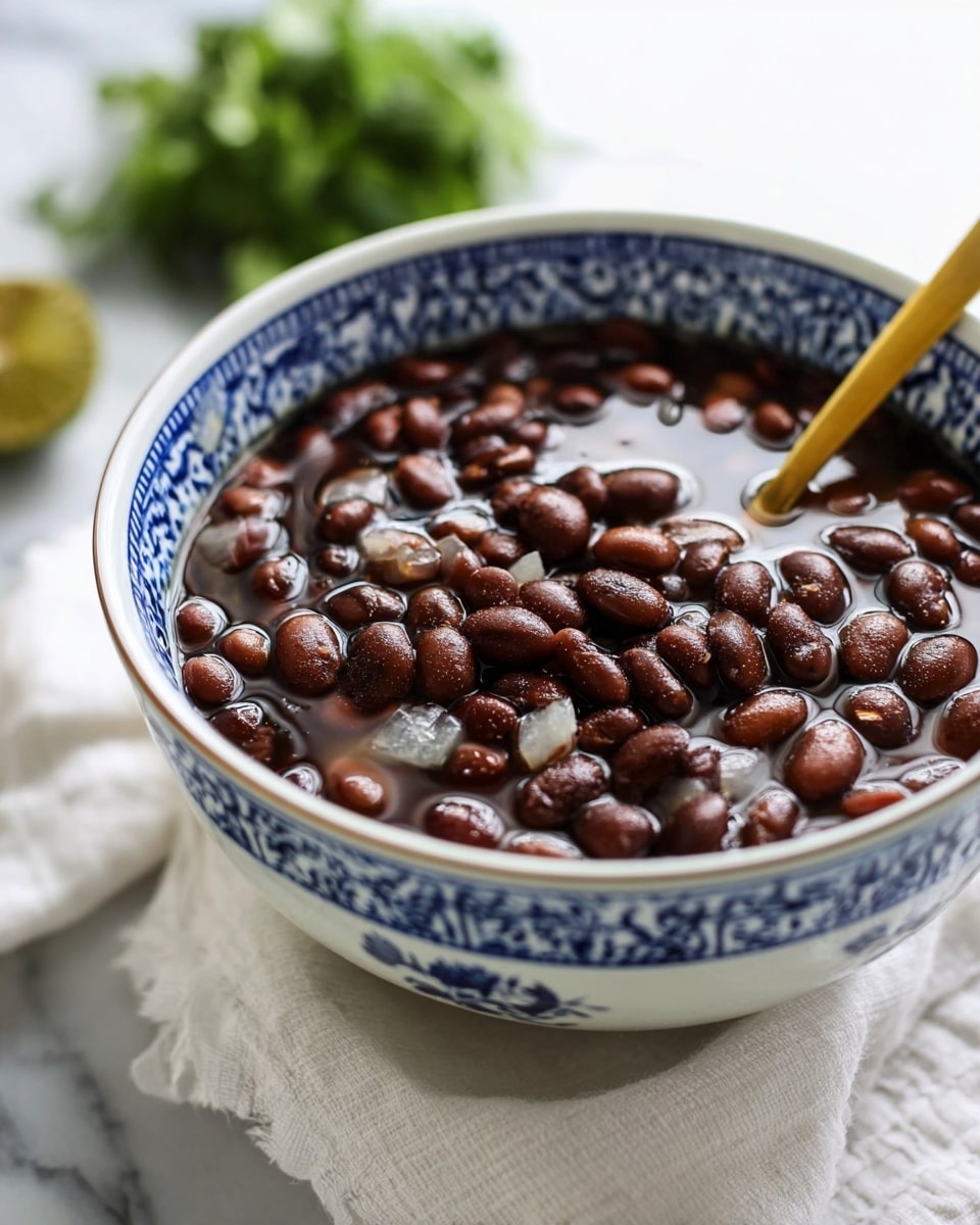 A close-up view of a white bowl with blue patterns filled with dark brown cooked beans mixed with small pieces of soft white onion in a thick liquid. A golden spoon lifted above the bowl is holding a spoonful of the beans and onions. In the background, there is a blurred green leafy garnish on a white marbled surface. The lighting is soft, highlighting the shiny, moist texture of the beans. photo taken with an iphone --ar 4:5 --v 7